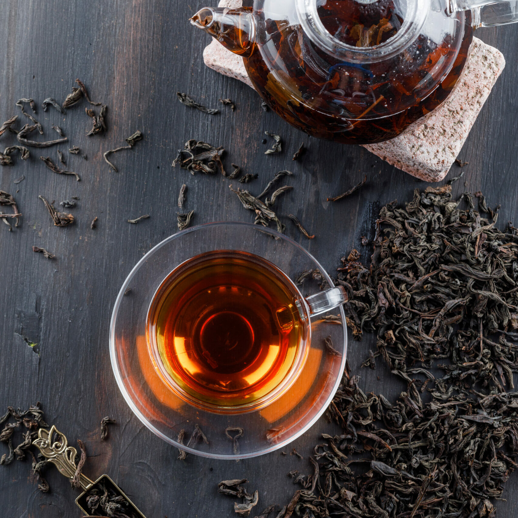 Black tea with dry tea, brick in teapot and cup on wooden background, top view.